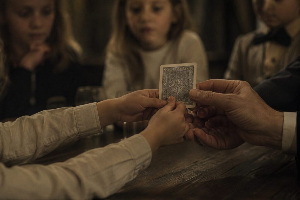 Zauberer reicht einem Kind eine Spielkarte, weitere Kinder verfolgen den Trick am Tisch