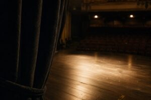 Blick aus der Seitengasse auf eine leere, beleuchtete Theaterbühne, im Vordergrund die Kante eines schweren Samtvorhangs
